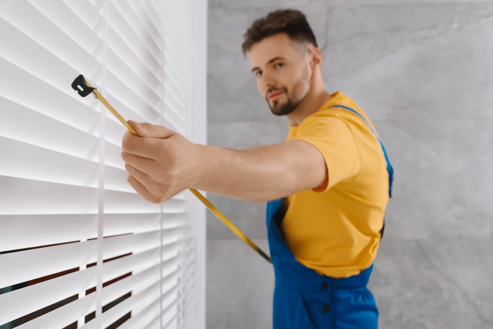 Un artisan menuisier en tenue professionnelle prenant des mesures précises d'une fenêtre intérieure ou d'une baie avec un mètre laser ou un décamètre, dans une maison résidentielle, montrant son professionnalisme et sa concentration sur les détails. La scène se déroule dans un intérieur clair et accueillant, avec un or deux clients présents, écoutant les explications de l'artisan, incarnant le suivi personnalisé et la relation directe entre le professionnel et le client, du diagnostic à la préparation de l'installation.