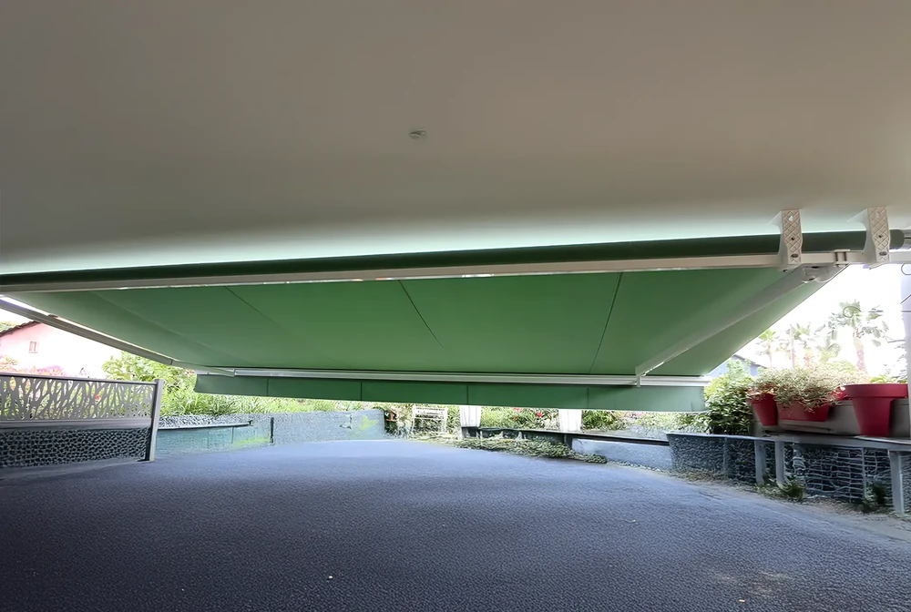 Terrasse pavée d'une villa provençale, vue de trois-quarts, avec un store extérieur de couleur taupe ou blanc cassé déployé protégeant une zone de détente. Le store montre clairement ses bras articulés et sa structure aluminium en configuration déployée. À proximité, un coin lounge avec fauteuils et table basse en rattan. En arrière-plan, végétation méditerranéenne et ciel bleu. La photo est prise en plein jour, par beau temps, mettant en évidence l'ombre nette créée par le store et le confort de l'espace protégé du soleil. L'atmosphère est résidentielle, chaleureuse et professionnelle.