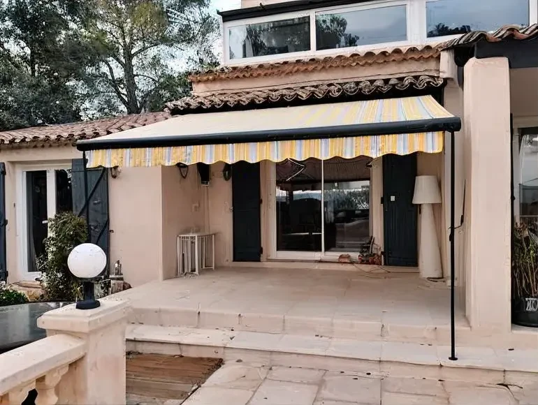 Maison individuelle de style méditerranéen ou provençal située dans l'Est-Var, vue de la façade principale en façade jour. La maison dispose d'une baie vitrée ou d'une large ouverture d'accès à une terrasse, équipée d'un store extérieur toile dépliée de couleur neutre (beige, gris ou blanc cassé) fournissant une ombre généreuse et bien définie sur la terrasse. Le store doit être clairement visible avec ses bras articulés et sa structure de fixation en aluminium. La terrasse en carrelage accueille du mobilier de détente (chaises longue ou salon de jardin), et quelques éléments de décoration (pots de fleurs, parasol optionnel en arrière-plan). La photo est prise en condition d'ensoleillement direct, par beau temps, mettant en évidence le contraste ombre/lumière et le confort apporté par le store. L'ambiance générale est résidentielle, professionnelle et accueillante, distincte de l'image déjà sélectionnée par sa perspective ou son environnement immédiat (jardin différent, architecture maison différente).